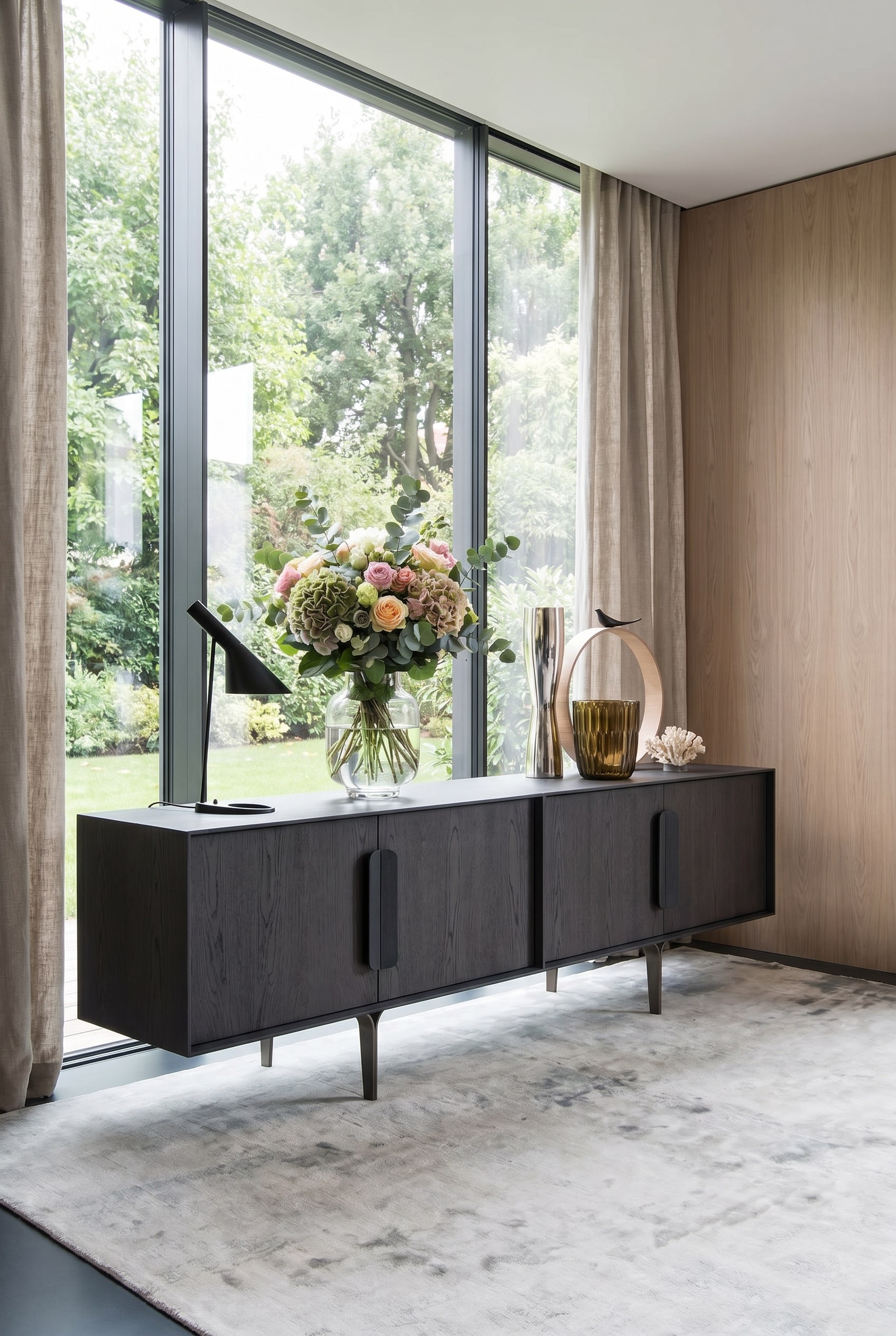 Dunkles Designer-Sideboard vor gro&szlig;en Fenstern, darauf eine helle, leuchtende Statement-Vase mit Fr&uuml;hlingsblumen