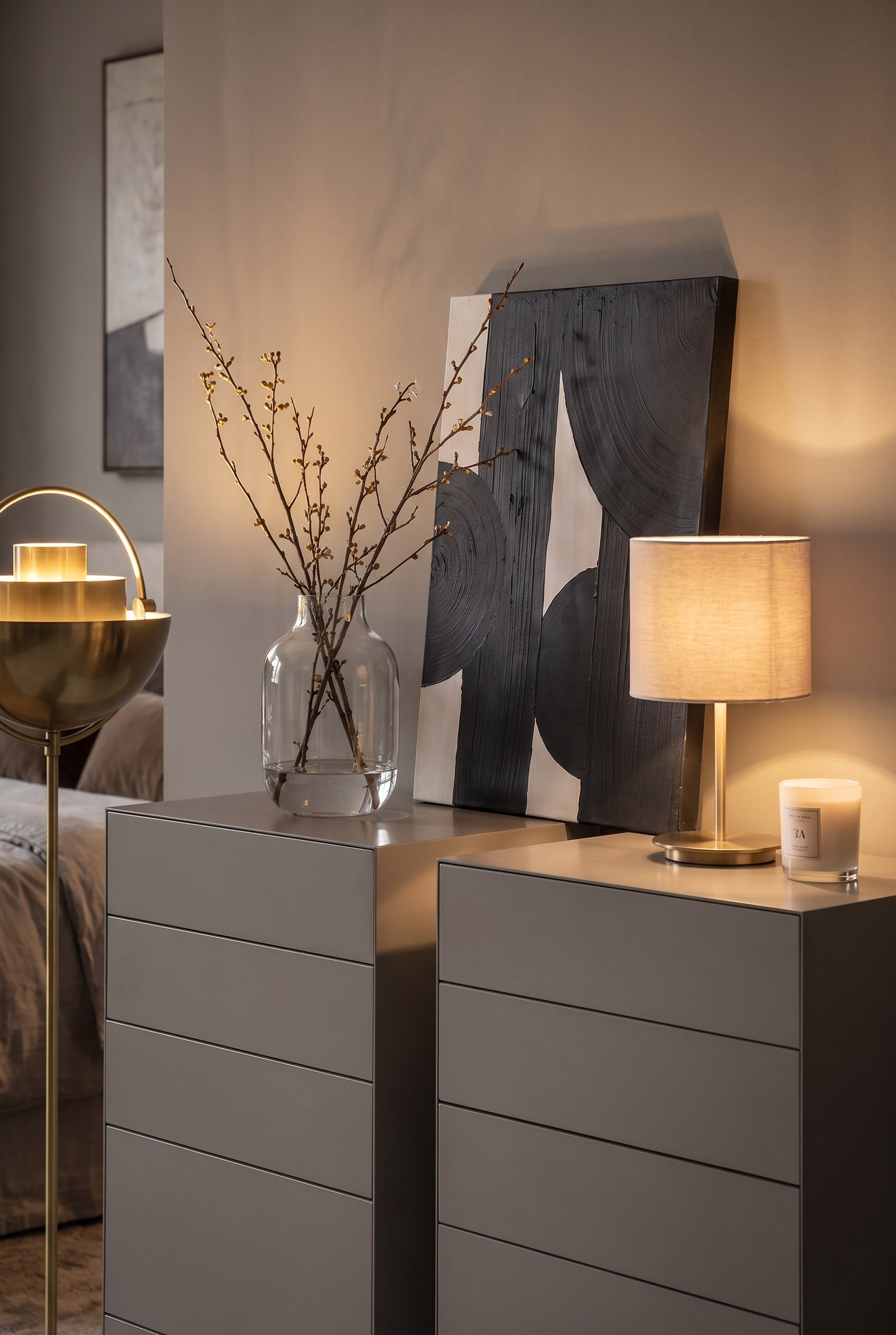 Decorazione su due cassettiere alte grigie: Un quadro astratto appoggiato, un vaso di vetro con rami e una lampada da tavolo.