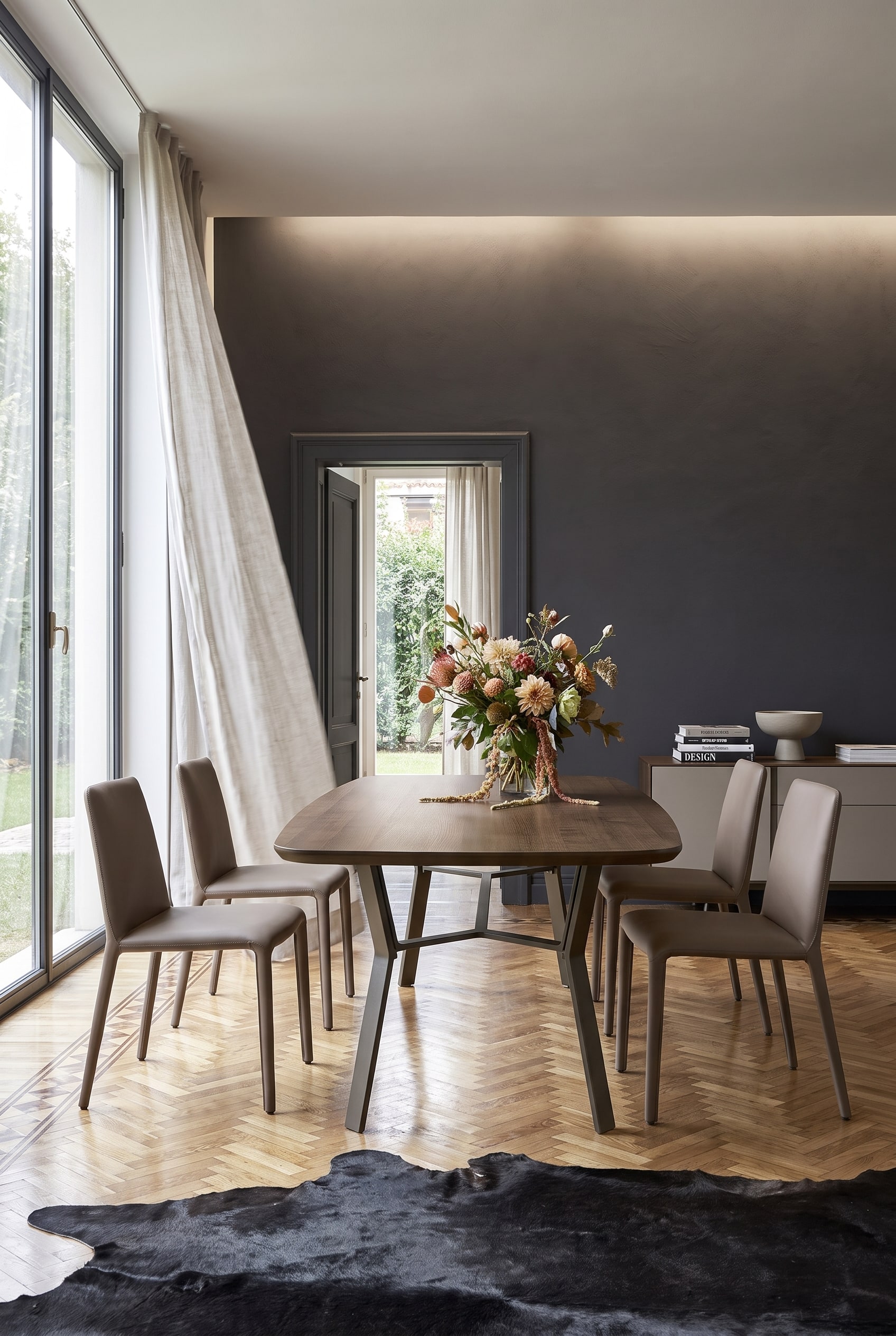 Sala da pranzo minimalista con parete accentata scura, tavolo in legno con base in ferro e quattro sedie in pelle color tortora