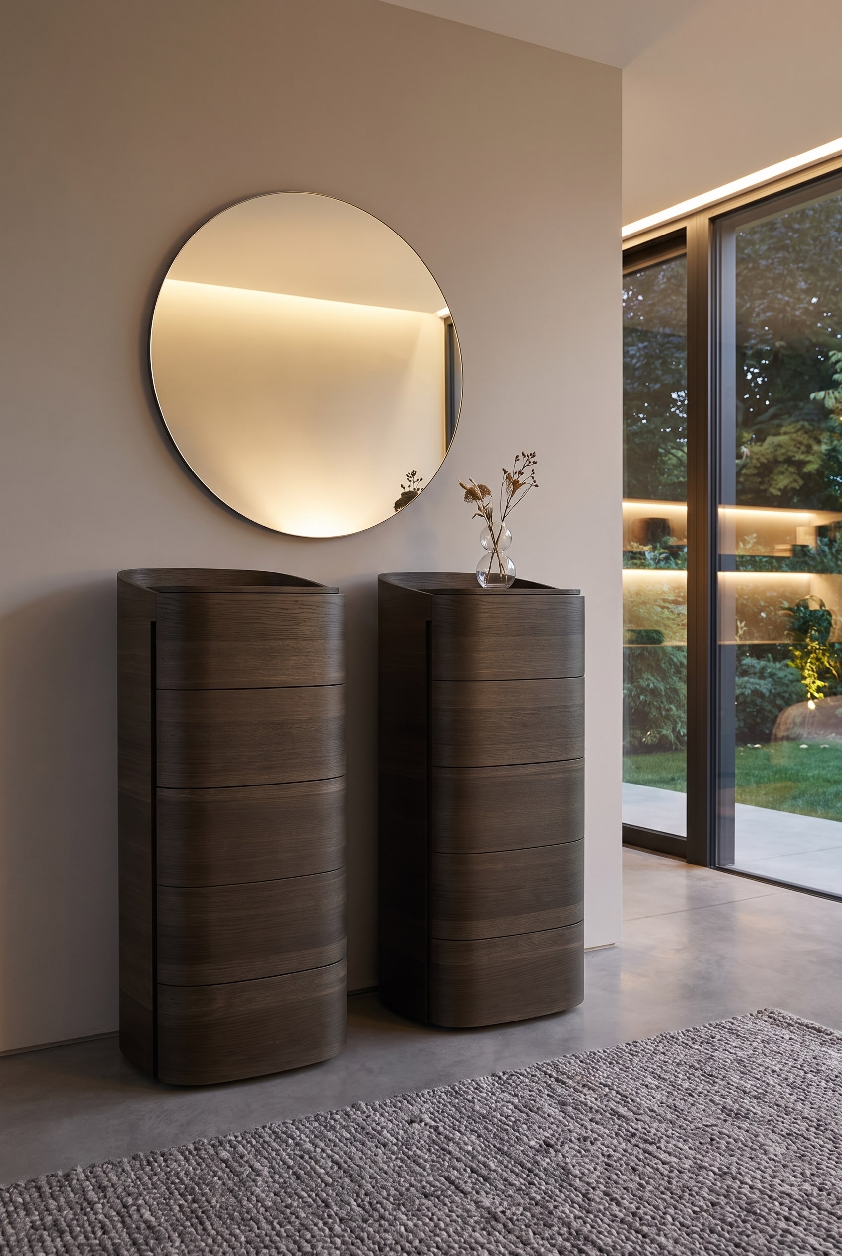 Tall, rounded chest of drawers made of dark wood, with a large, minimalist round mirror on the wall above it, and a small vase in front