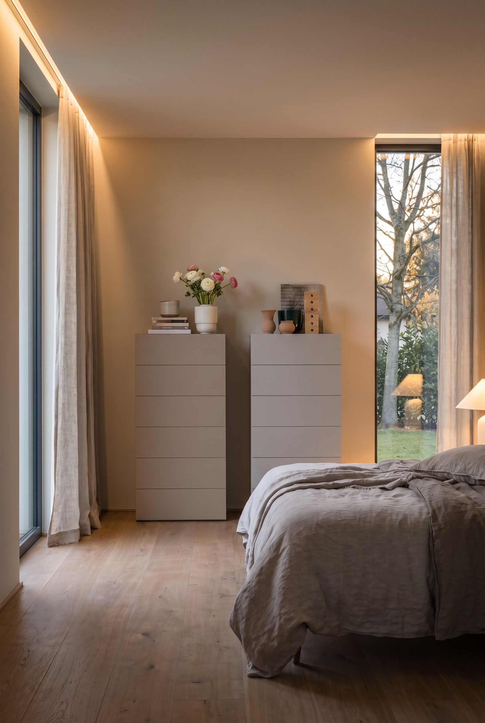 Twee greige hoge ladekasten in een moderne slaapkamer met linnen beddengoed en kamerhoge ramen.