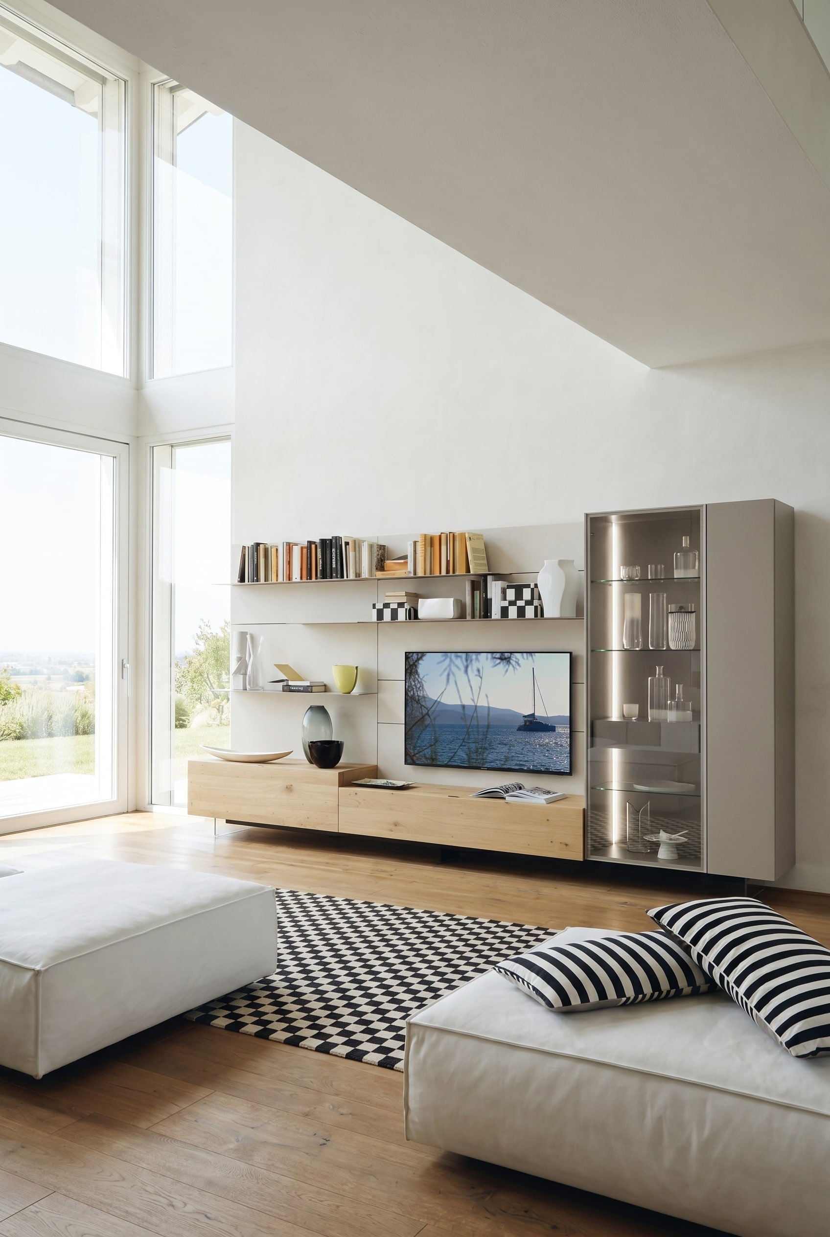 Soggiorno luminoso con moduli base sospesi in rovere chiaro, mensole bianche e una vetrina alta, illuminata con luce calda