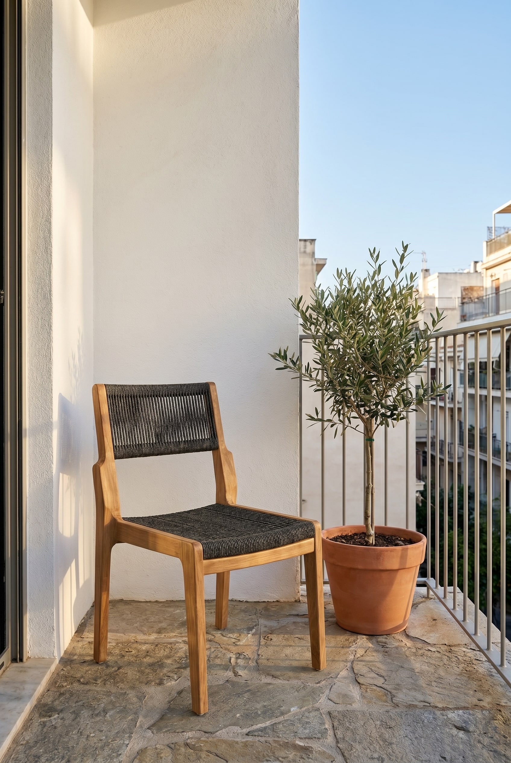 Balcone compatto con pavimento in pietra naturale e una sedia in legno di teak con intreccio in corda color antracite nella dolce luce del mattino