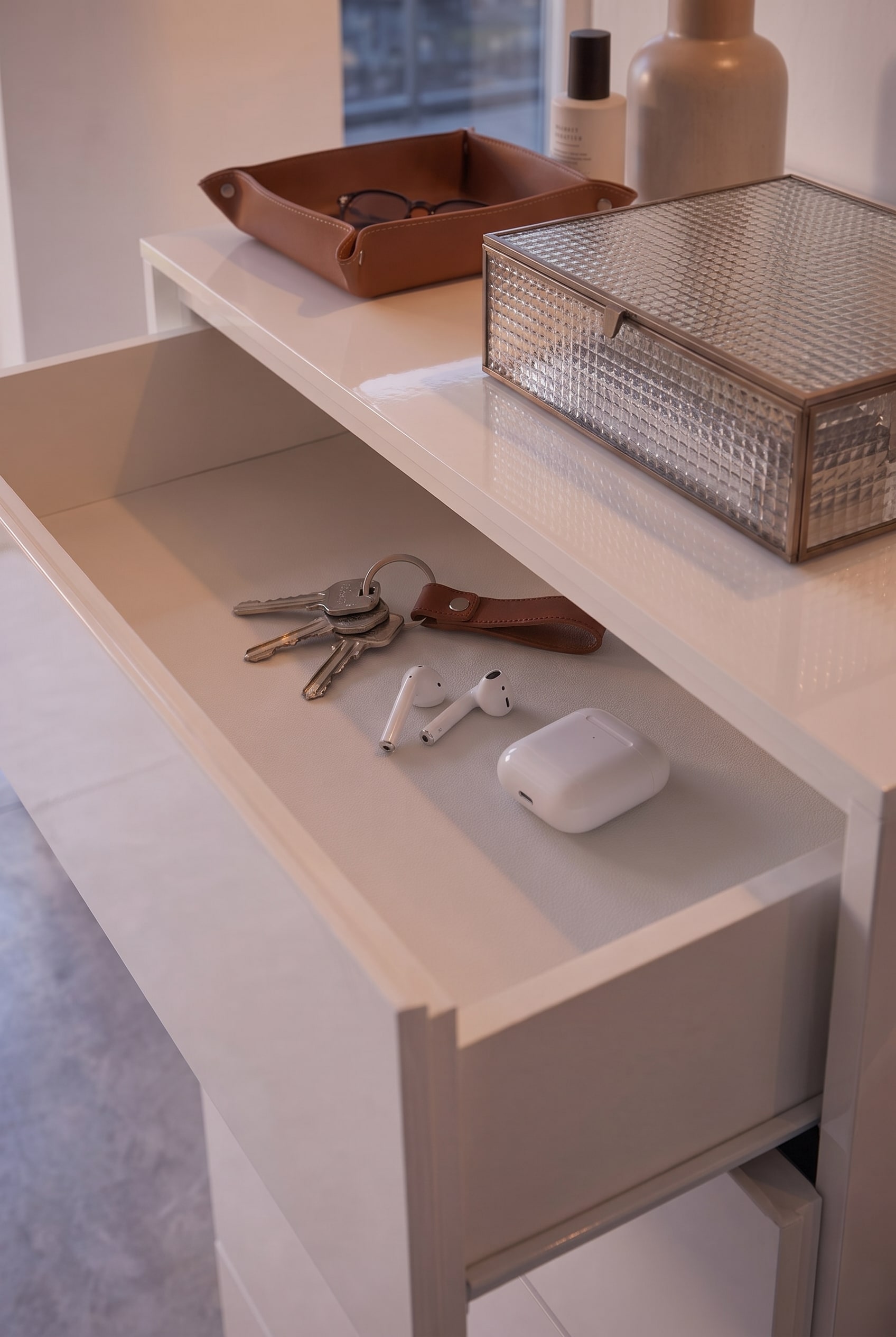 Cassetto piatto aperto di una cassettiera bianca, al suo interno sono disposti in modo ordinato chiavi, AirPods e un portachiavi in pelle.