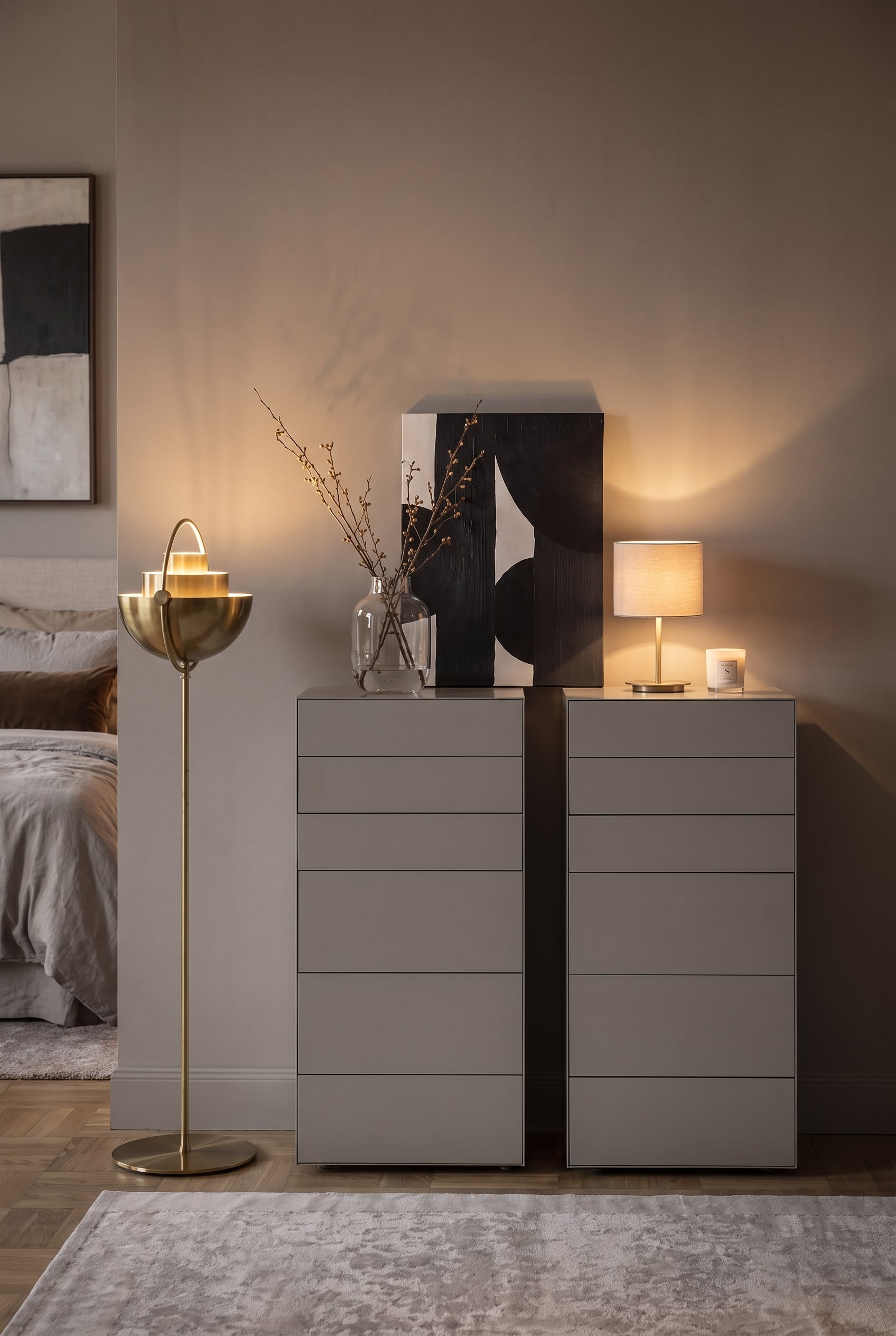 Vista frontale di due cassettiere alte grigie con decorazione asimmetrica e una lampada da terra dorata a sinistra.