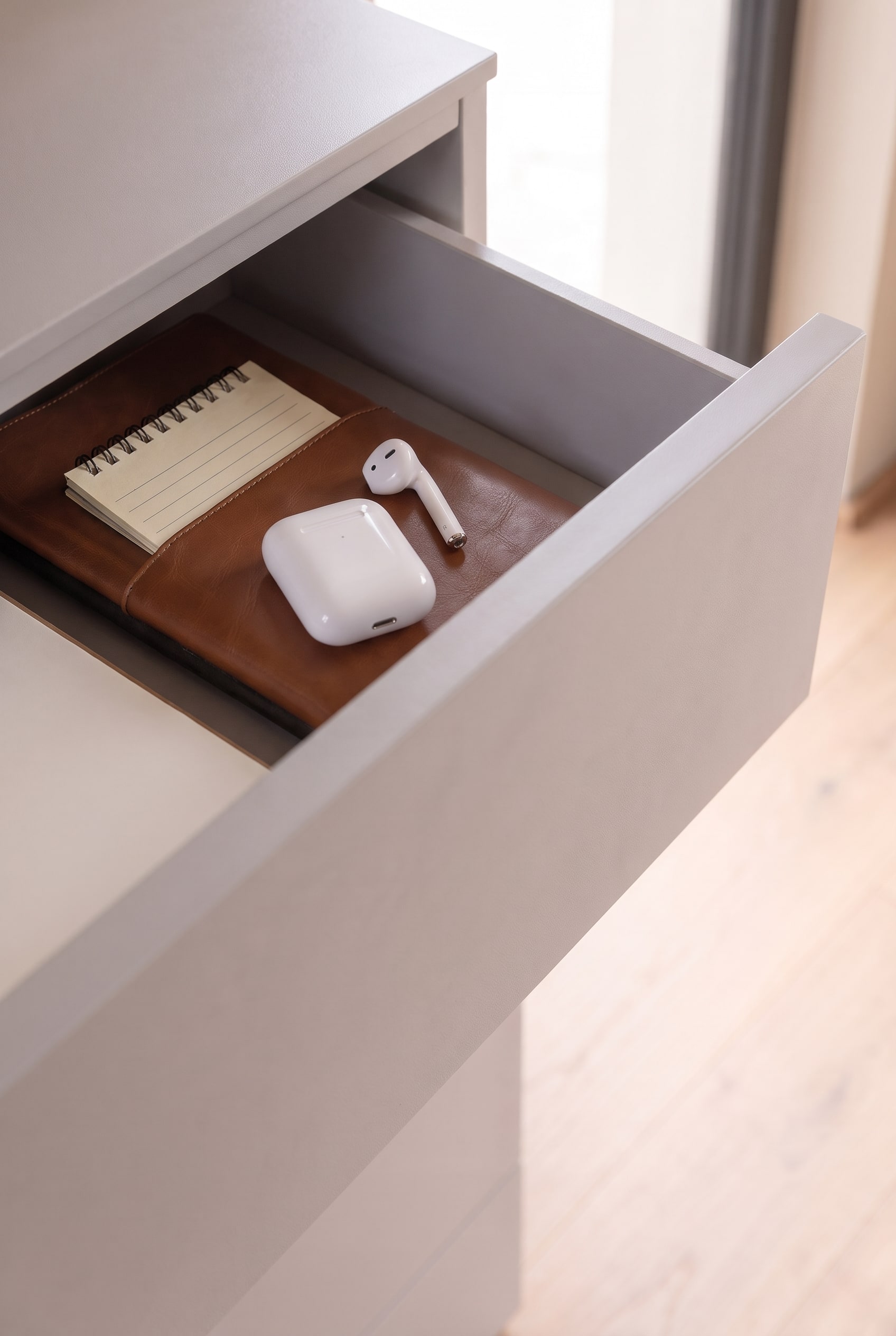 Geopende lade van een greige ladekast met een leren notitieboek en witte AirPods.